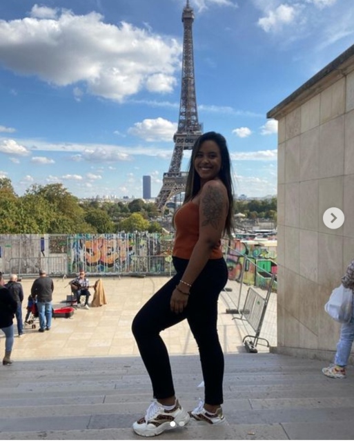 Foto da filhota, numa escada de calça preta e blusa marron, com a torre Eiffel ao fundo. Ela sorri para a foto.